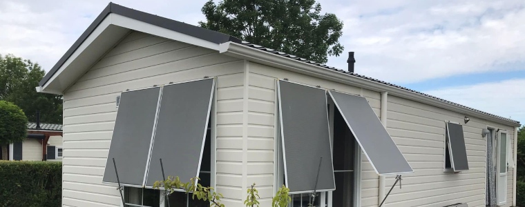 Is een chalet waardevast? Een chalet met witte gevelbekleding, grijze zonneschermen en een zadeldak, omgeven door groene struiken en bomen.