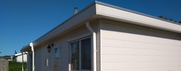 Hoe lang gaat een chalet mee? Een gerenoveerd chalet met witte gevelbekleding, een plat dak en witte dakgoten onder een blauwe lucht.